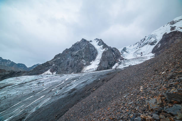 istock Dramatic landscape with glacier tongue and icefall on large snow mountain range with sharp rocky pinnacle under gray cloudy sky. Long glacier in high altitude. Gloomy scenery in mountains in overcast. 1562844808