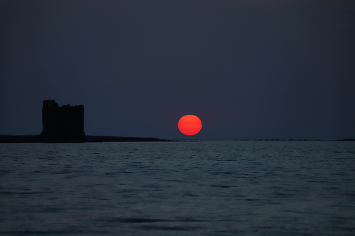 Vibrant Blood Orange coloured Sun setting to the right of  Torre della Finanza in Silhouette. Seen sailing through the Fornelli Channel looking out to Sea over the Isola Piana, Province of Sassari, Sardinia