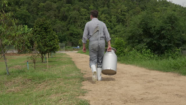 A male dairy farmer walking outside his farm holding a large milk can