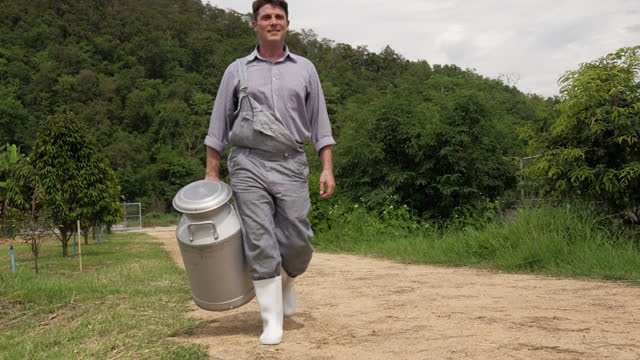 A male dairy farmer walking outside his farm holding a large milk can