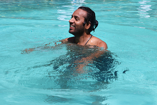Stock photo showing close-up view of swimming pool with turquoise blue mosaic tiles lining the length of the pool.  Indian man swimming, splashing and playing in crystal clear water.