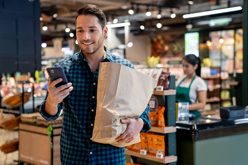 Portrait of a Latin American man leaving the supermarket carrying a shopping bag and checking his new account balance on his cell phone