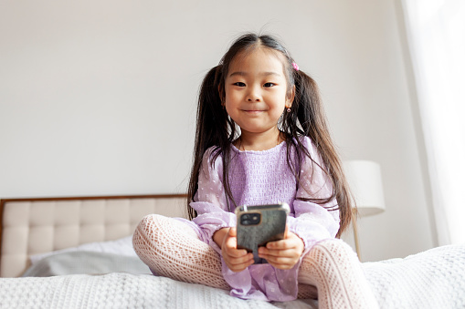 little asian girl is sitting on the bed at home and using smartphone, the child is looking at the phone and typing message and smiling in purple dress