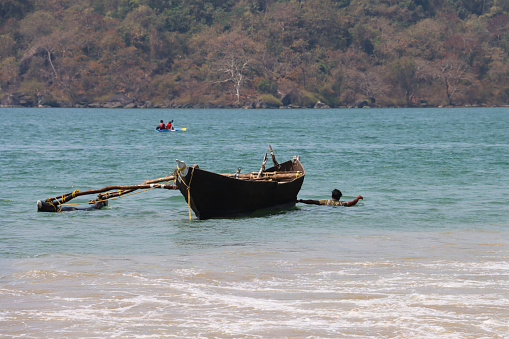 Palolem Beach, Goa, India - March, 7 2023: Stock photo showing close-up view of boat floating in bay of water in Goa, India being dragged nearer the beach by a fisherman shoulder deep in the sea.
