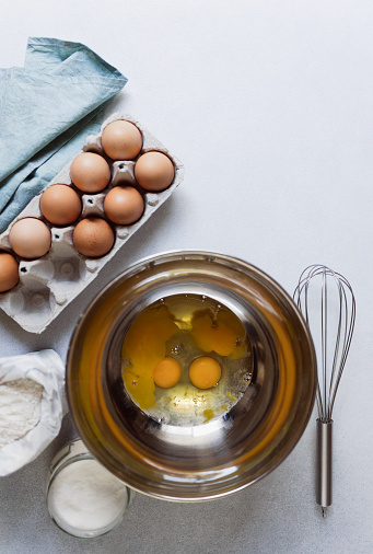 Raw eggs in stainless bowl. Carton of eggs, flour, sugar and kitchen textile napkin on concrete background. Ingredients for baking cake. Vertical photo, copy space