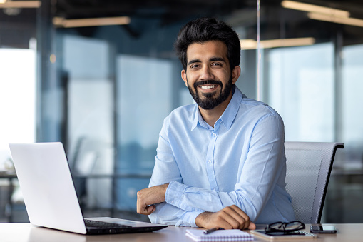 Portrait of happy and successful businessman, indian man smiling and looking at camera, satisfied with achievement results man working inside office building using laptop at work.