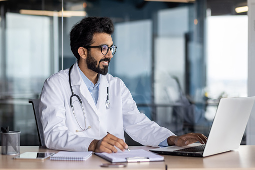 Successful smiling doctor working inside modern clinic office, hindu man in white medical coat working in medical office sitting at table with laptop.