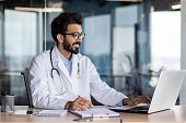 Successful smiling doctor working inside modern clinic office, hindu man in white medical coat working in medical office sitting at table with laptop