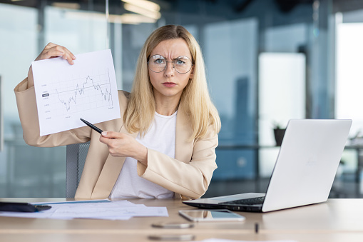 Upset and worried young woman manager, business owner sitting in office in front of computer and showing documents. Online business meeting, reporting.