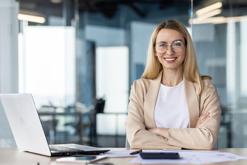 Portrait of a successful and self-confident business woman in a business suit, sitting in the office at the desk and looking at the camera, smiling with her arms crossed.