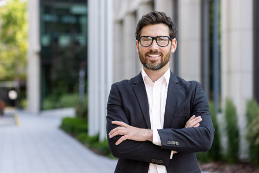 Portrait of mature adult businessman, senior boss with beard smiling and looking at camera, experienced and successful man in business suit outside office building with crossed arms.