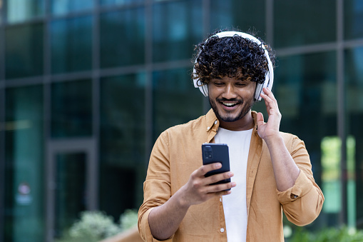 A young male Hispanic student is standing on the street near the campus wearing headphones with a phone in his hands. Listens to music, a podcast, talks on a video call.