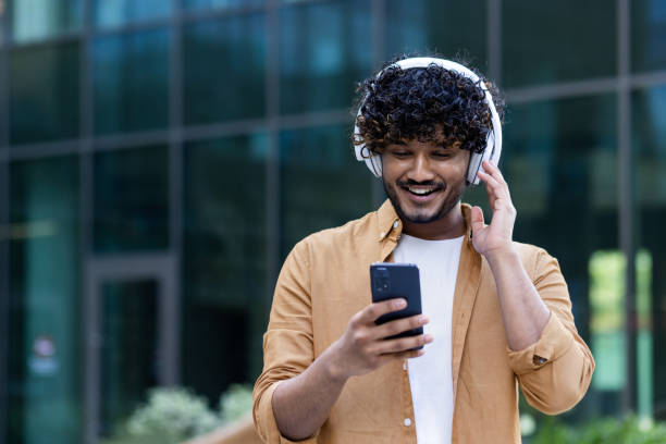 a young male hispanic student is standing on the street near the campus wearing headphones with a phone in his hands. listens to music, a podcast, talks on a video call - indian ethnicity audio imagens e fotografias de stock