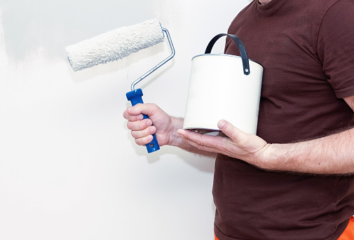 A man with a bucket of paint in his hand and a paint roller against the background of a wall and paint stains.