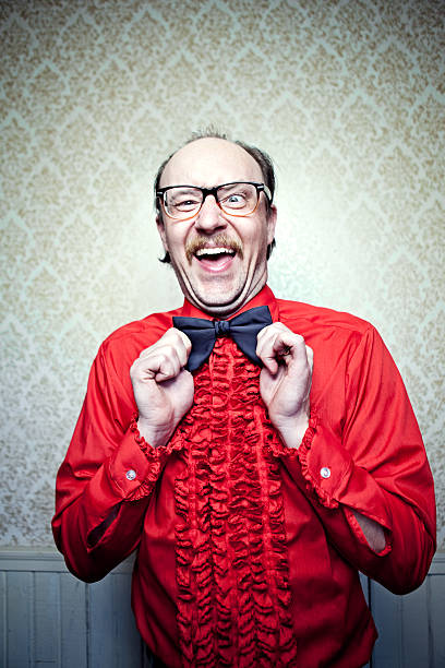 A balding man in a retro frilly red tuxedo shirt, mustache, and glasses, makes a goofy face while straightening his bow tie.  Vintage damask pattern wallpaper on wall in the background.  Vertical with copy space.