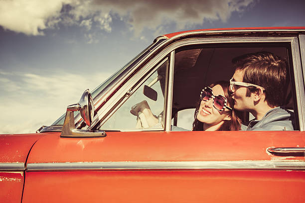 Car Ride Couple Retro A young man kisses his girlfriend wearing white sunglasses, visible through the driver window of their classic 1960s car.  Vintage styling and colors.  Wide and low angle to emphasis the blue sky and clouds.  Horizontal with copy space. city break photos stock pictures, royalty-free photos & images