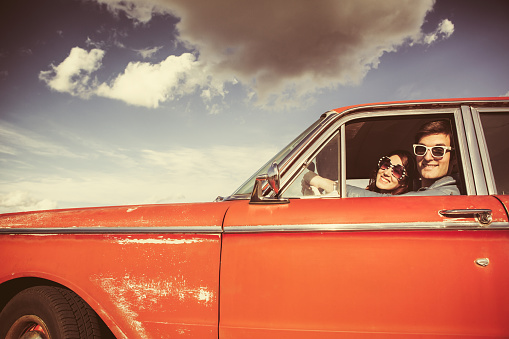 An attractive young man and woman wearing white sunglasses smile out the driver window as they drive in their classic 1960s car.  Vintage styling and colors.  Wide and low angle to emphasis the blue sky and clouds.  Horizontal with copy space.