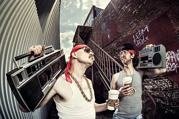 Two stereotypical hip hop / gangsta / rapper characters with baggy pants, gold chains (bling), and doo rags stand in a graffiti covered urban city alley.  Both are holding stereo cassette players (boom box) on their shoulders, and humorously enjoying to-go cups of coffee while having a deep discussion.  High contrast gritty post process.  Horizontal with copy space.