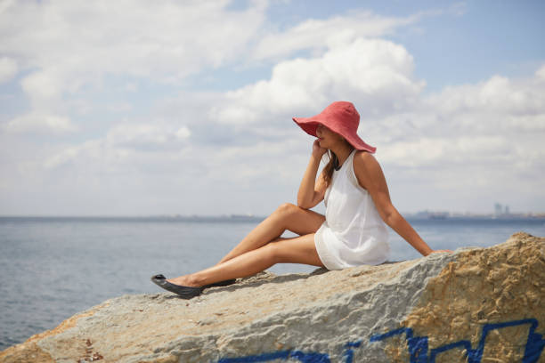 Beautiful Kazakh tourist girl with huge red hat sitting over rocks near lighthouse at Istanbul, with amazing old city in background