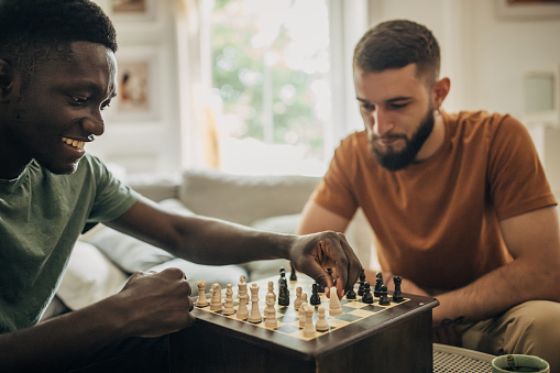Two people, male friends playing a game of chess in living room at home.