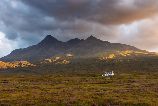 A small white cottage in the remote wilderness near Sligachan on the Isle of Skye, Scotland, UK. A dramatic sky can be seen alongside the golden hour evening light with a stunning view of the Cuillin mountain range in the background.