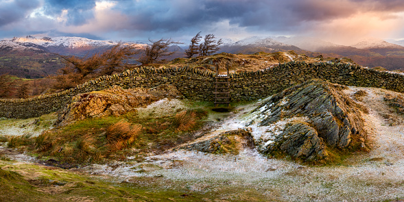 Rural stone wall and wooden stile with dramatic Winter panoramic view of snowcapped mountain range. Black Fell, Lake District, UK.