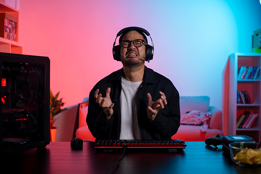 Angry young Asian man playing a video game feeling upset after loosing