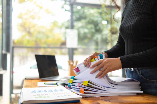 secretary searches through stacked documents on desk in office to find lease within stacked documents just before meeting. concept difficulty in finding hire purchase contract from stacked documents
