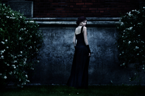 Low key portrait of a creepy woman in black dress standing against stone wall.