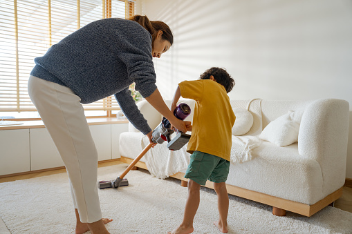 Asian mother teaching her son uses a vacuum cleaner for cleaning the living room on weekends.