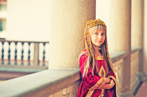 Full-length portrait of little girl dressed like medieval person in old-fashioned dress with accessories posing against vintage background. Concept of historical remake, comparison of eras, fashion.