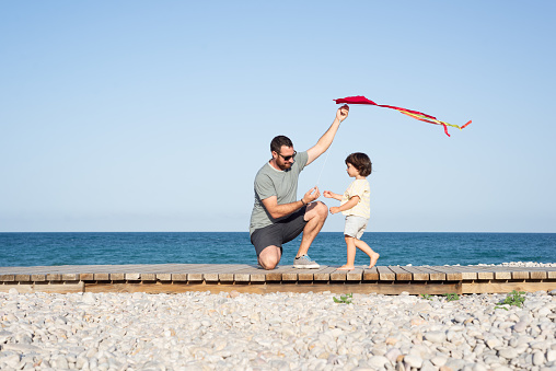 Father is teaching her little daughter to fly a kite at a mediterranean pebble beach.