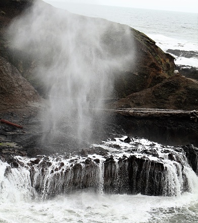 an incoming wave has sent a spray of water and mist upwards while water also rushes over the hard rock which forms The Spouting Horn at Cape Perpetua.