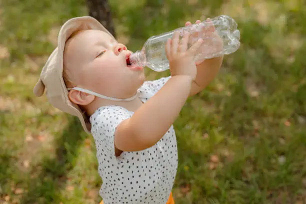 Photo of Thirst, dehydration. Cute little boy drinks water from a plastic bottle against on the playground. Portrait of a thirsty handsome boy. The benefits of drinking water.