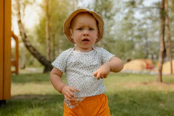 Photo of Thirst, dehydration. Cute little boy drinks water from a plastic bottle against on the playground. Portrait of a thirsty handsome boy. The benefits of drinking water.