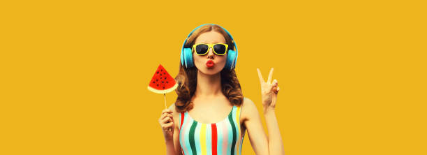 Summer portrait of stylish woman in headphones listening to music blowing her lips with juicy lollipop or ice cream shaped slice watermelon on yellow background stock photo