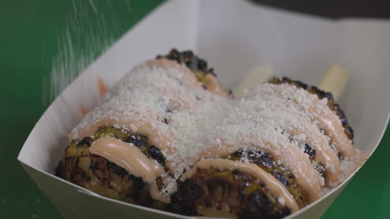 Chef preparing elote corn sprinkling cheese on grilled corn, close-up