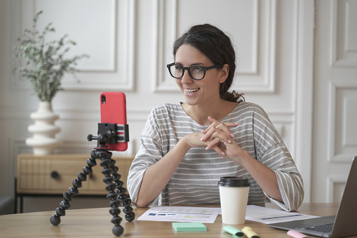 Smiling young spanish businesswoman in glasses sitting in front of smartphone on tripod at workplace, positive female influencer recording video for blog, sharing skills while shooting online training