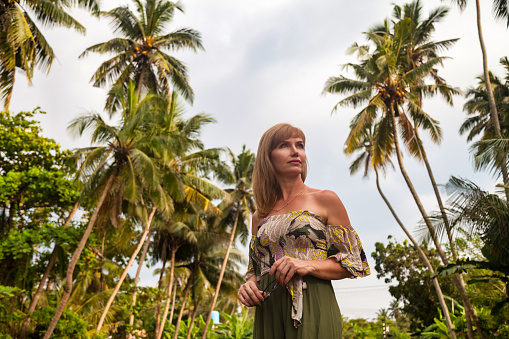 Middle aged lady at tropical jungles background, looking away. Smiling adult woman 50 year old in nature Sri Lanka in tropics with green palm trees. Travel summer vacation concept. Copy ad text space