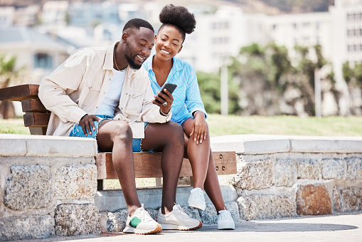 Black couple, love and phone while browsing social media, internet or watching a video while sitting on a park bench. Happy man and woman on a romantic date, trip or summer holiday to relax together