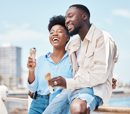 Happy couple at the beach eating an ice cream cone while on a date in nature while on spring vacation. Black man and woman talking and laughing while hugging and having dessert on holiday by seaside.