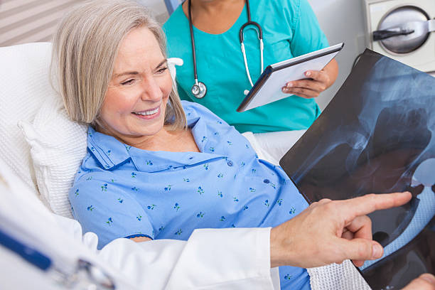 Happy Senior Woman Patient in Hospital Bed Happy senior woman patient recovering in hospital bed with male doctor and female nurse looking at hip replacement x-ray hip replacement stock pictures, royalty-free photos & images