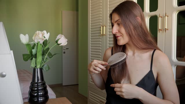 Smiling brunette woman sitting at makeup table and brushing her long hair. Hair care, female elegance and beauty