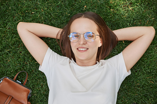 Young Caucasian woman lying on grass with hands behind head wearing white t shirt and eyeglasses, enjoying summertime and resting in nature.