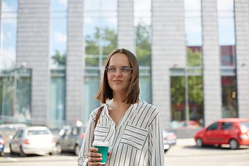 Outdoor portrait of beautiful young woman with brown hair holding cup of coffee walking in city street wearing white blouse and stylish eyeglasses.