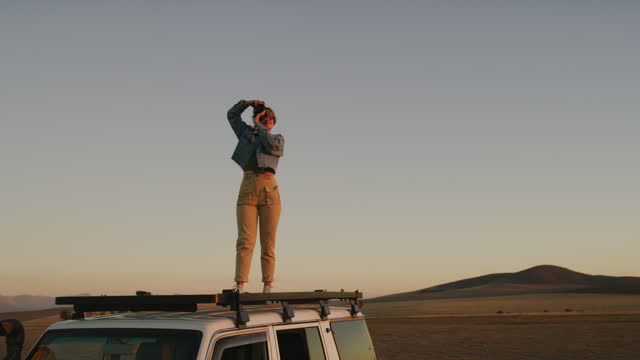 Travel, woman with camera on roof of car and nature photography at sunset, desert and trekking adventure. Road trip, photographer standing on vehicle and taking pictures of safari view in evening.