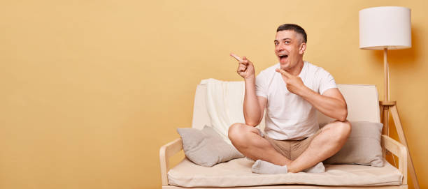 взволнованный смеющийся брюнет, молодой человек в повседневной белой футболке, сидящий на диване у бежевой стены, указывающий на рекламную - space to left стоковые фото и изображения
