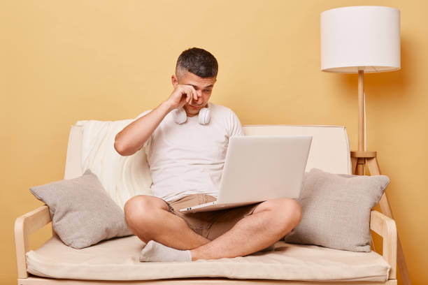 istock Freelancer lifestyle. Digital work. Online browsing. Exhausted young man working on laptop computer sitting on comfortable couch at home felling fatigue rubbing painful eyes 1563658225