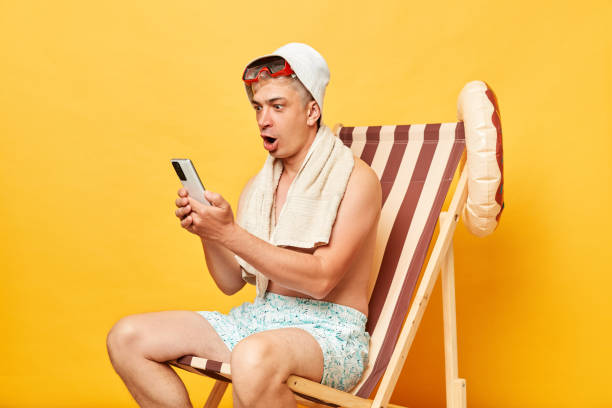 choqué effrayé jeune touriste en vêtements d’été et panama assis sur une chaise longue isolé sur fond jaune à l’aide d’un téléphone portable lisant un mauvais message. - panama deck beach chair photos et images de collection