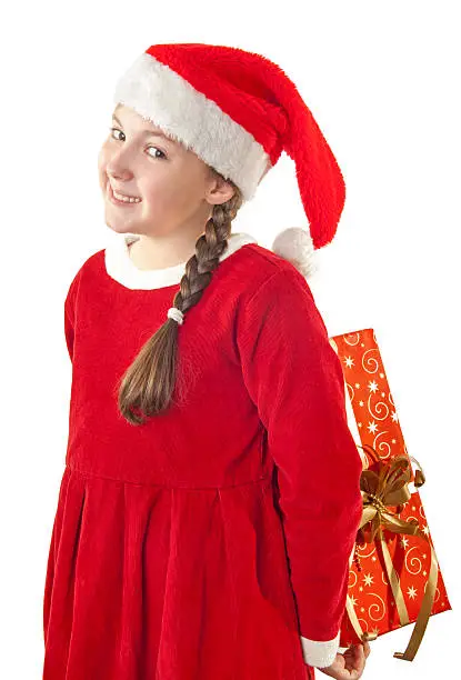 Beautiful girl dressed in Christmas clothes and red santa hat with present behind her back isolated on white background